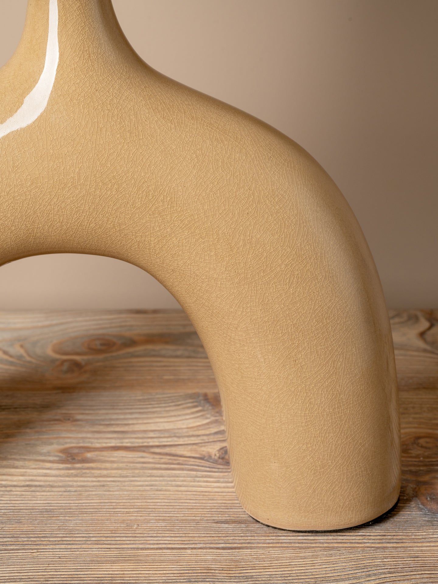 Close-up of a sculptural ceramic vase with a curved, organic shape in a warm sandy beige color, resting on a rustic wooden surface against a neutral beige background.