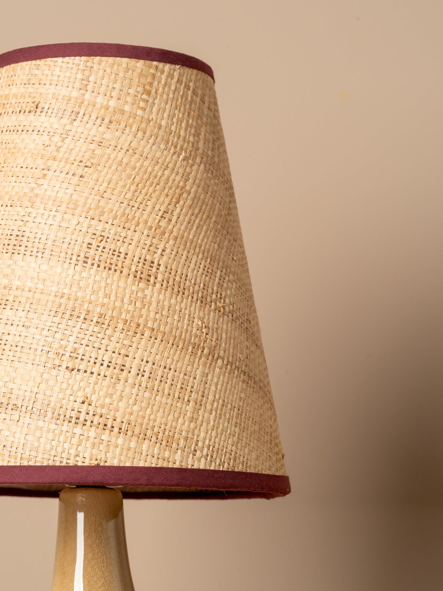 Close-up of a woven natural fiber lampshade with dark burgundy trim, mounted on a beige ceramic lamp base against a solid beige background.