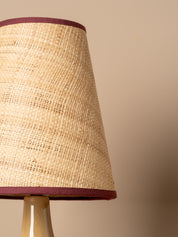 Close-up of a woven natural fiber lampshade with dark burgundy trim, mounted on a beige ceramic lamp base against a solid beige background.