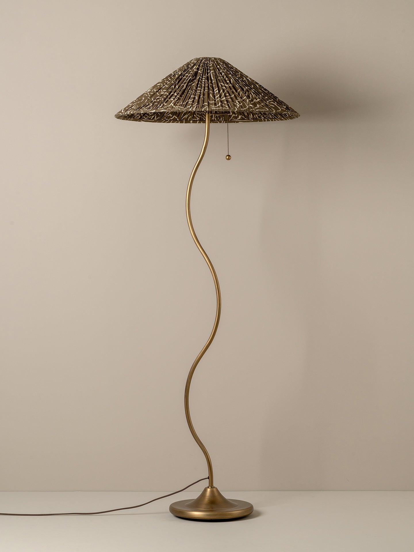 A vintage-style floor lamp with a wavy brass-colored metal stem, a round base, and a textured woven shade, shown against a plain beige wall.