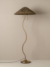 A vintage-style floor lamp with a wavy brass-colored metal stem, a round base, and a textured woven shade, shown against a plain beige wall.