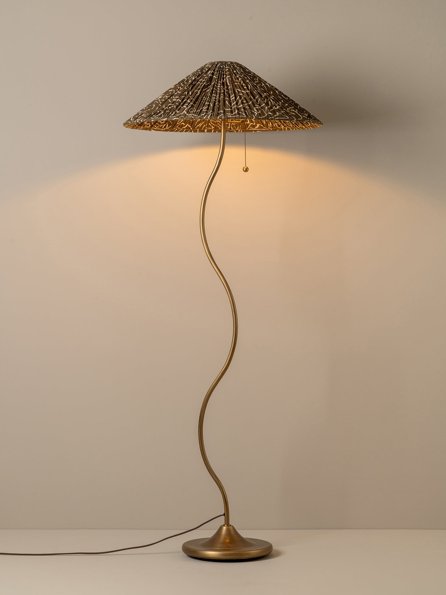 A mushroom-shaped floor lamp with a woven reed or straw conical shade, a curved metallic stem, and a round base, shown lit against a plain light beige wall.