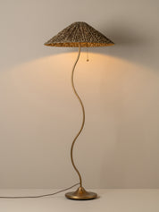 A mushroom-shaped floor lamp with a woven reed or straw conical shade, a curved metallic stem, and a round base, shown lit against a plain light beige wall.