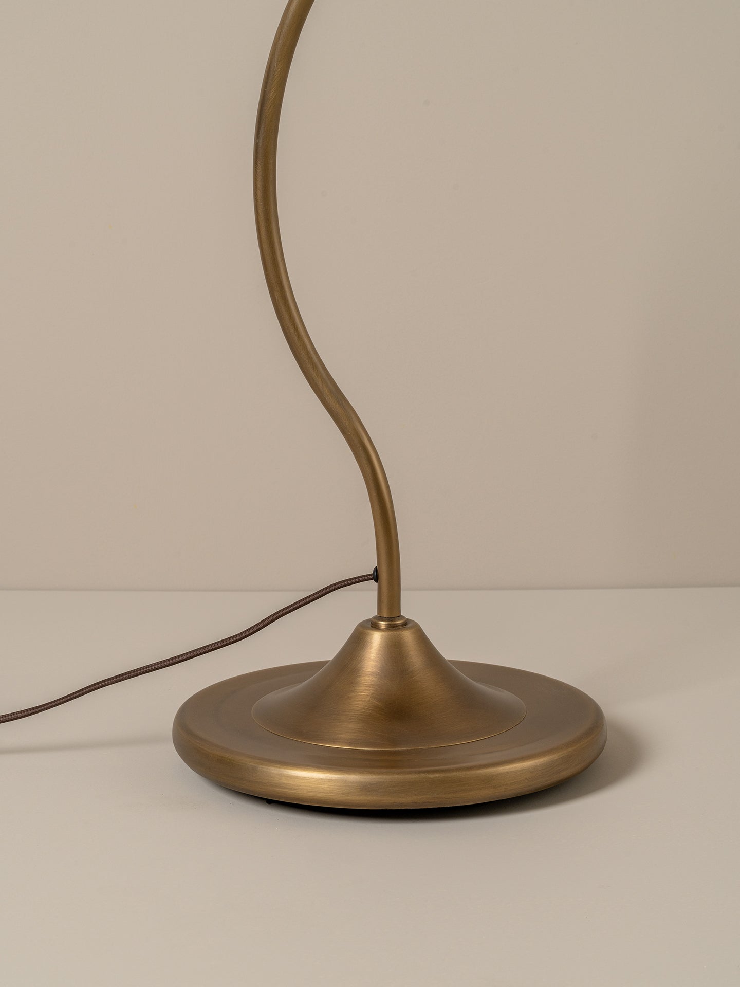 A close-up of a table lamp with a round brushed brass base and a curved flexible neck, shown against a neutral beige background.