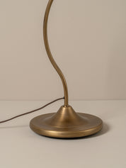 A close-up of a table lamp with a round brushed brass base and a curved flexible neck, shown against a neutral beige background.
