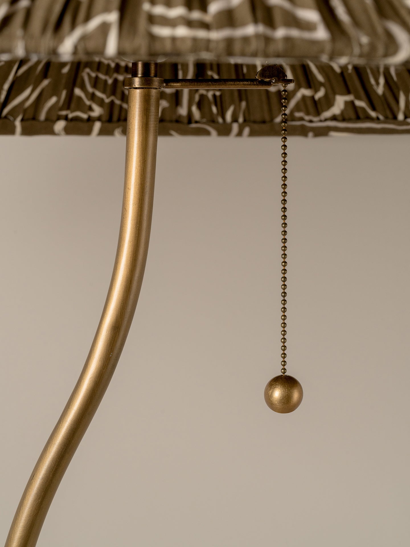 Close-up of a lamp with a curved brass pull chain and a patterned fabric shade against a neutral background.