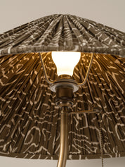 Close-up of a pleated fabric lampshade with an abstract brown and beige pattern, illuminated by a warm glowing bulb on a brass-toned metal fixture.