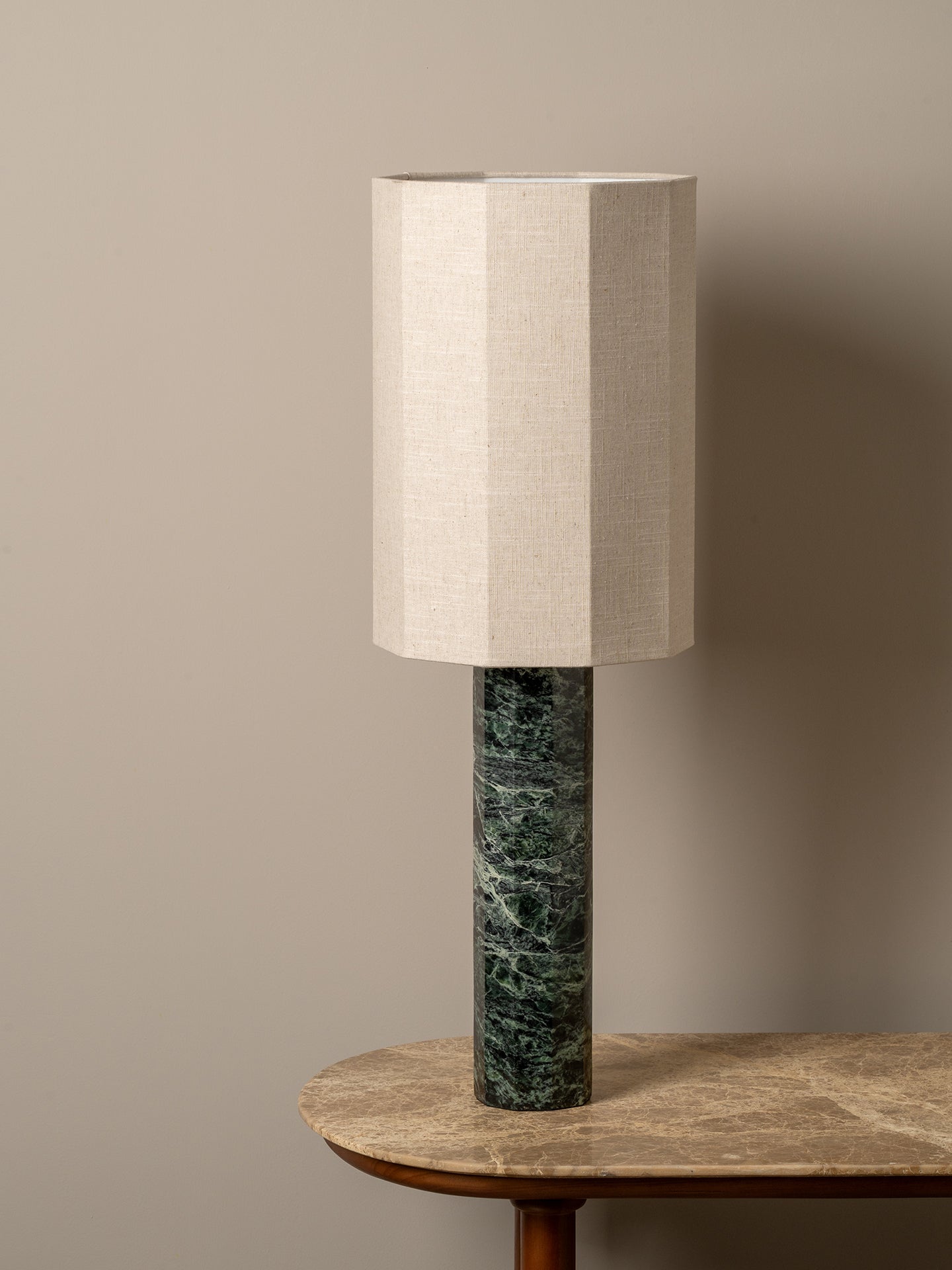 A table lamp with a beige linen drum shade and a dark green marble cylindrical base, displayed on a wooden table against a neutral beige wall.