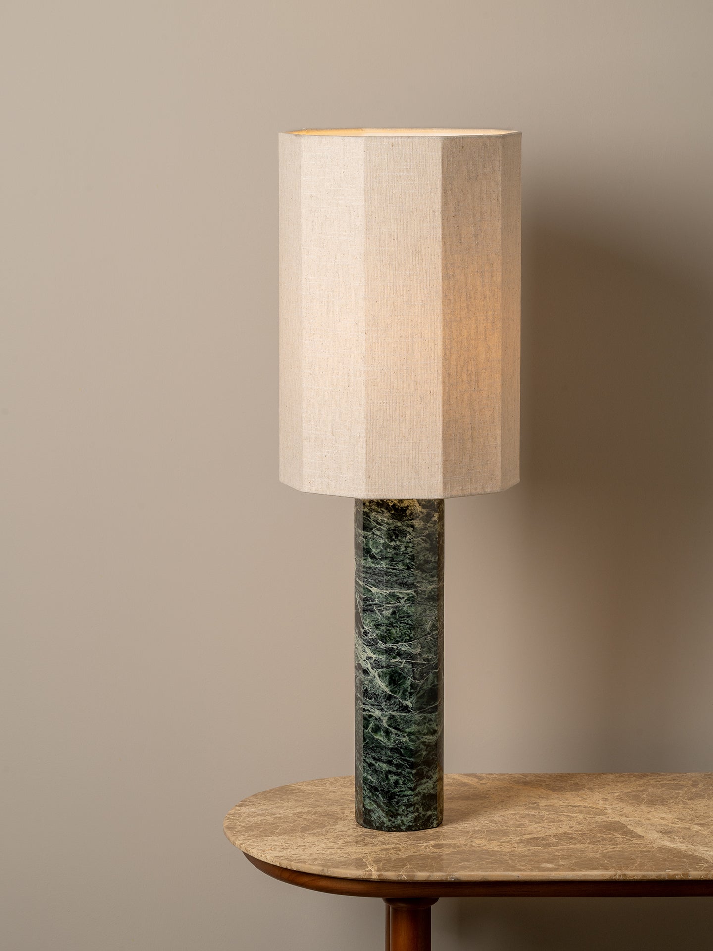 A table lamp with a beige fabric drum shade and a dark green marble base, shown lit on a wooden table against a neutral beige wall.