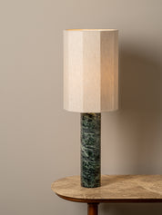 A table lamp with a beige fabric drum shade and a dark green marble base, shown lit on a wooden table against a neutral beige wall.
