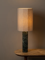 A table lamp with a warm beige linen drum shade and a dark green marble base, shown lit on a wooden table against a neutral brown background.