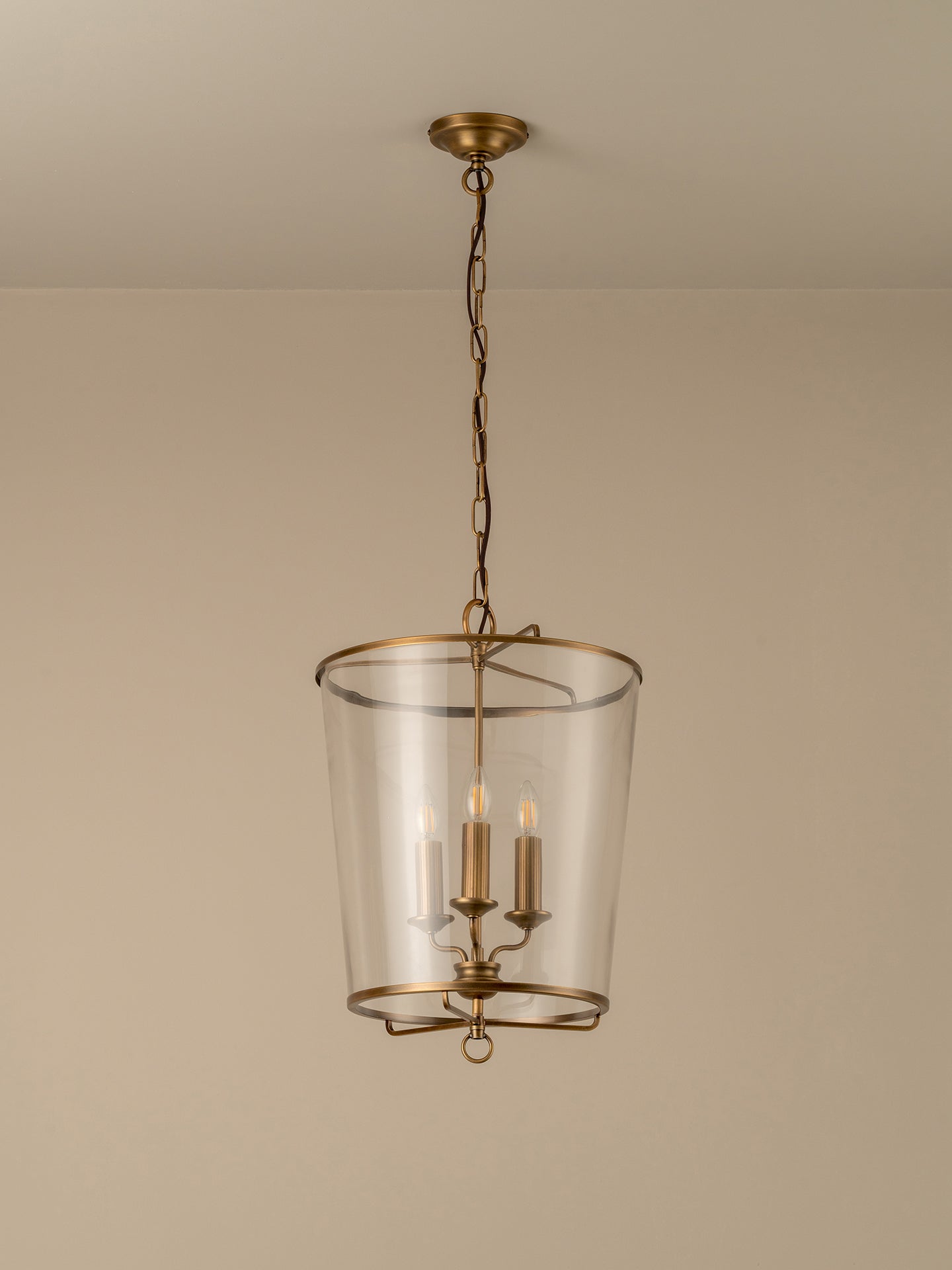 A hanging pendant light fixture with a clear glass cylinder shade and an antique brass or bronze metal frame, suspended by a chain from a matching ceiling canopy against a plain beige wall.