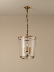 A hanging pendant light fixture with a clear glass cylinder shade and an antique brass or bronze metal frame, suspended by a chain from a matching ceiling canopy against a plain beige wall.