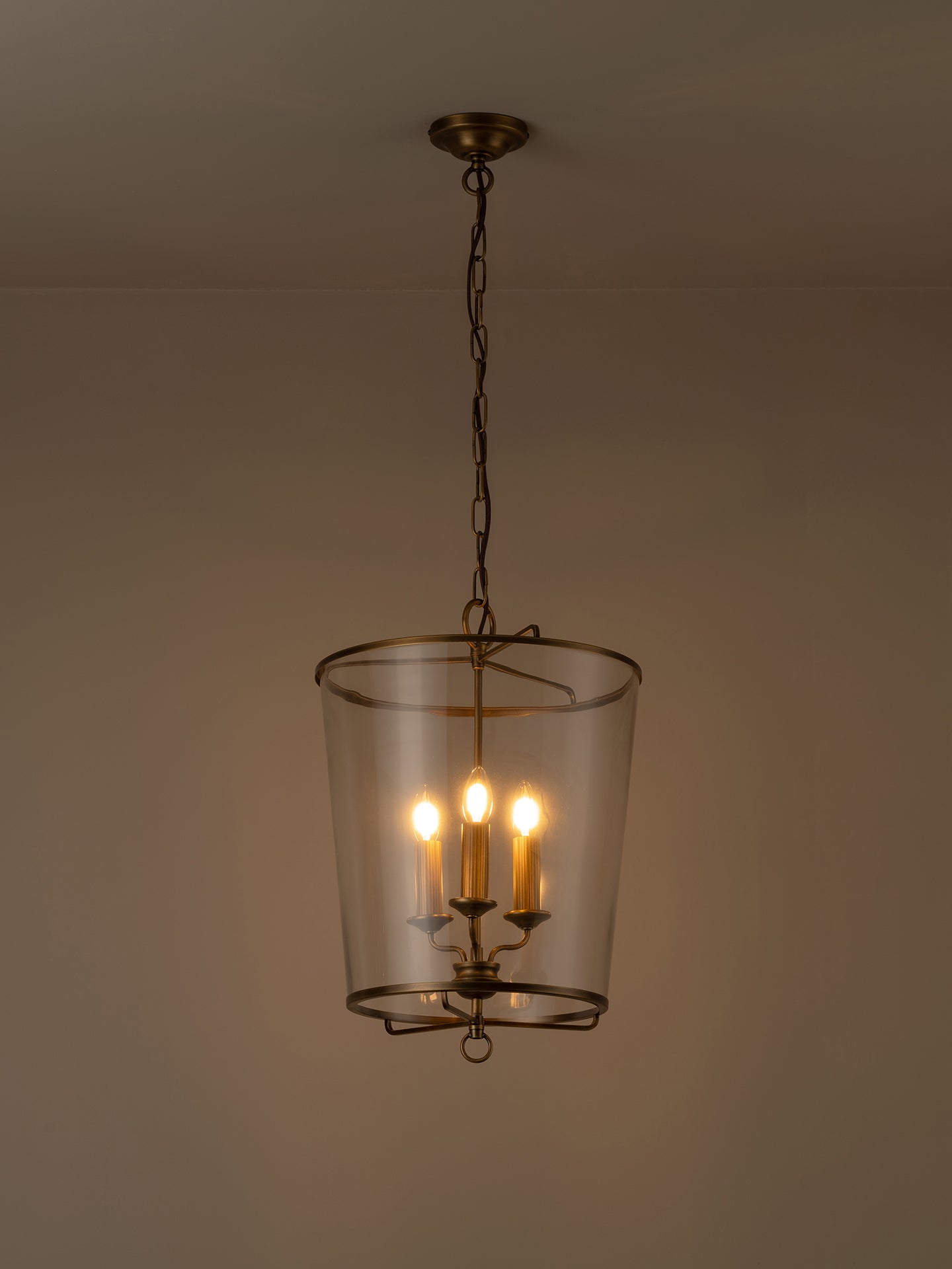 A hanging pendant light with a clear glass cylinder shade and warm brass-toned metal frame, suspended from a chain against a plain beige wall.