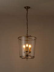 A hanging pendant light with a clear glass cylinder shade and warm brass-toned metal frame, suspended from a chain against a plain beige wall.