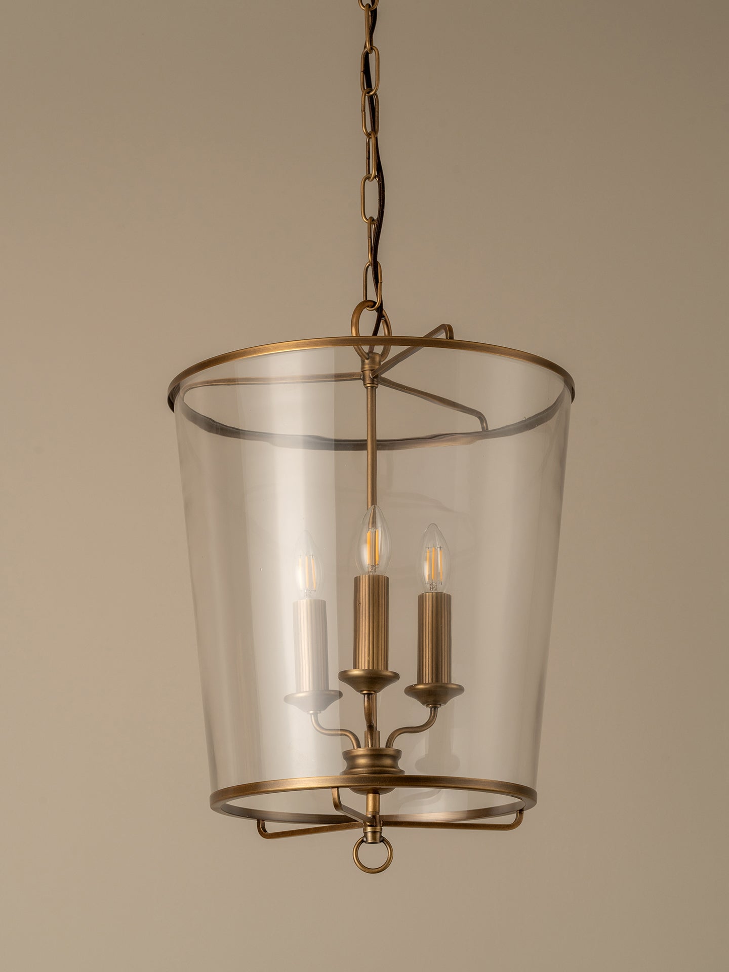 A hanging lantern-style pendant light with a clear glass cylindrical shade and antique brass metal frame and chain, shown against a plain warm beige background.