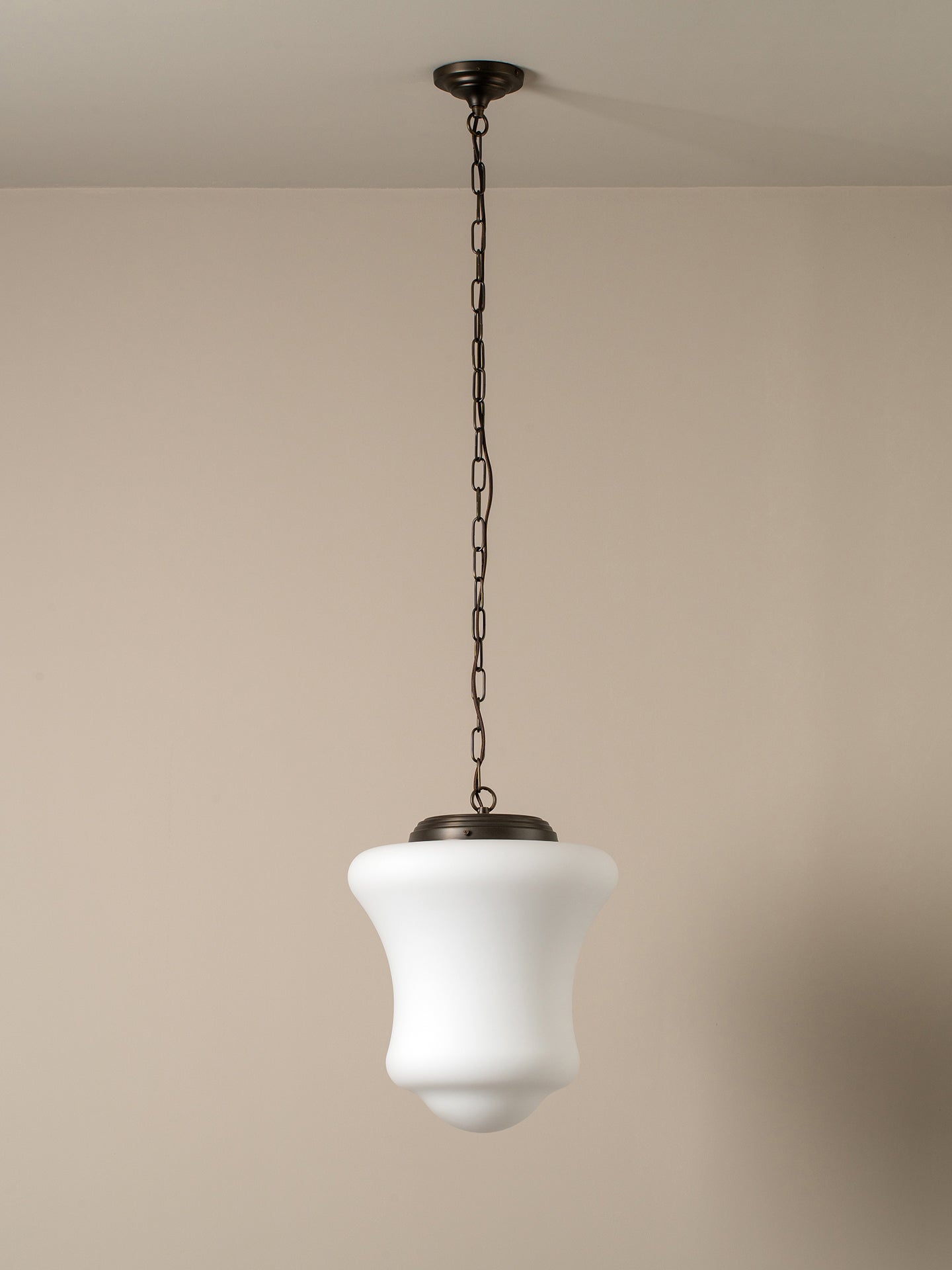 A modern pendant light with a white bell-shaped glass shade and a dark bronze metal canopy and chain, shown hanging against a neutral beige wall.