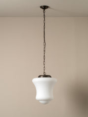 A modern pendant light with a white bell-shaped glass shade and a dark bronze metal canopy and chain, shown hanging against a neutral beige wall.