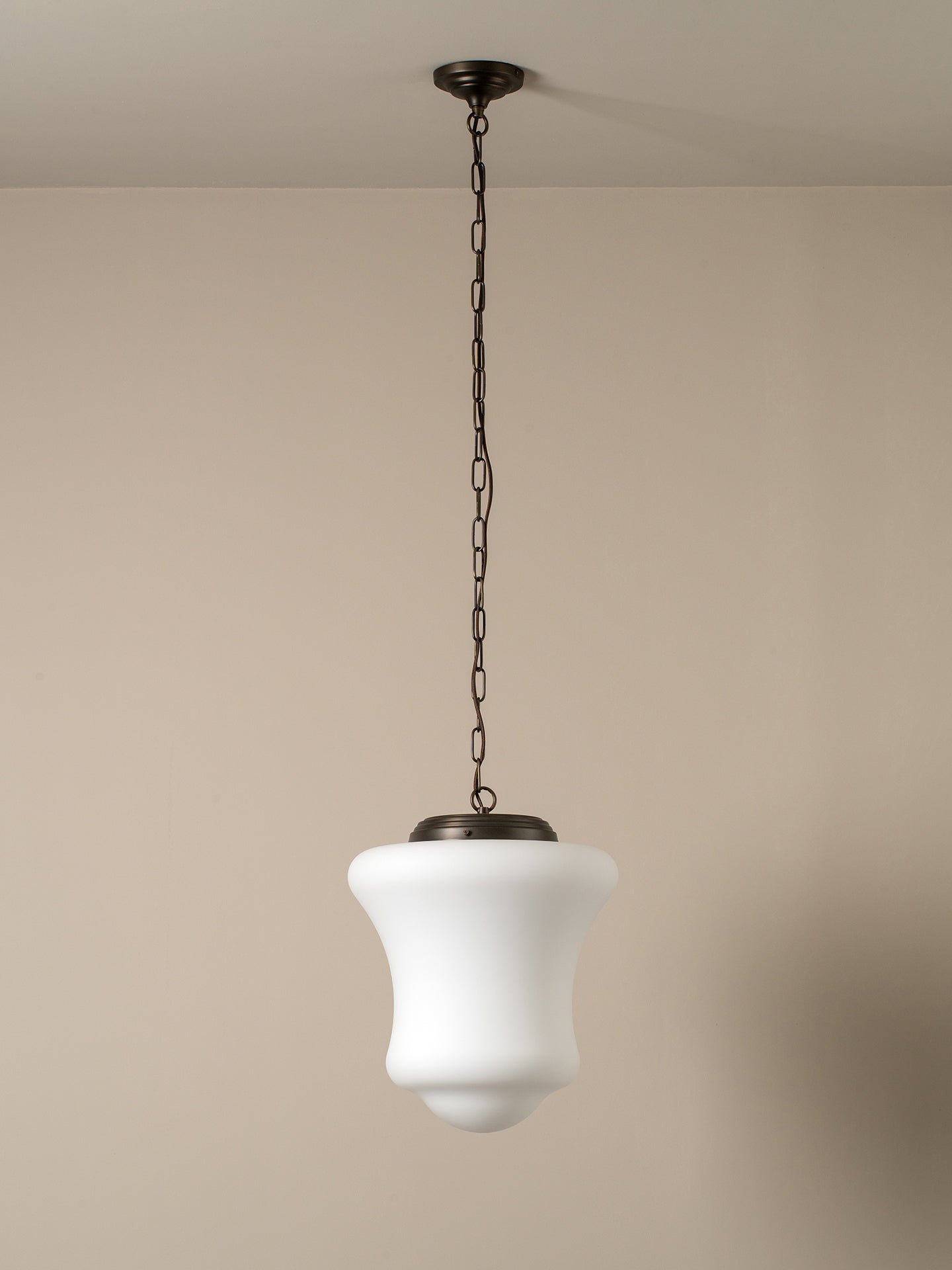 A modern pendant light with a white bell-shaped glass shade and a dark bronze metal canopy and chain, shown hanging against a neutral beige wall.
