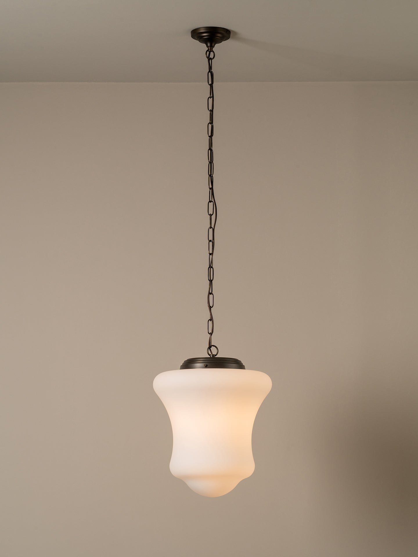 A single pendant light with a black metal chain and canopy, featuring a flared frosted glass shade that glows warm white, shown hanging against a plain beige wall.