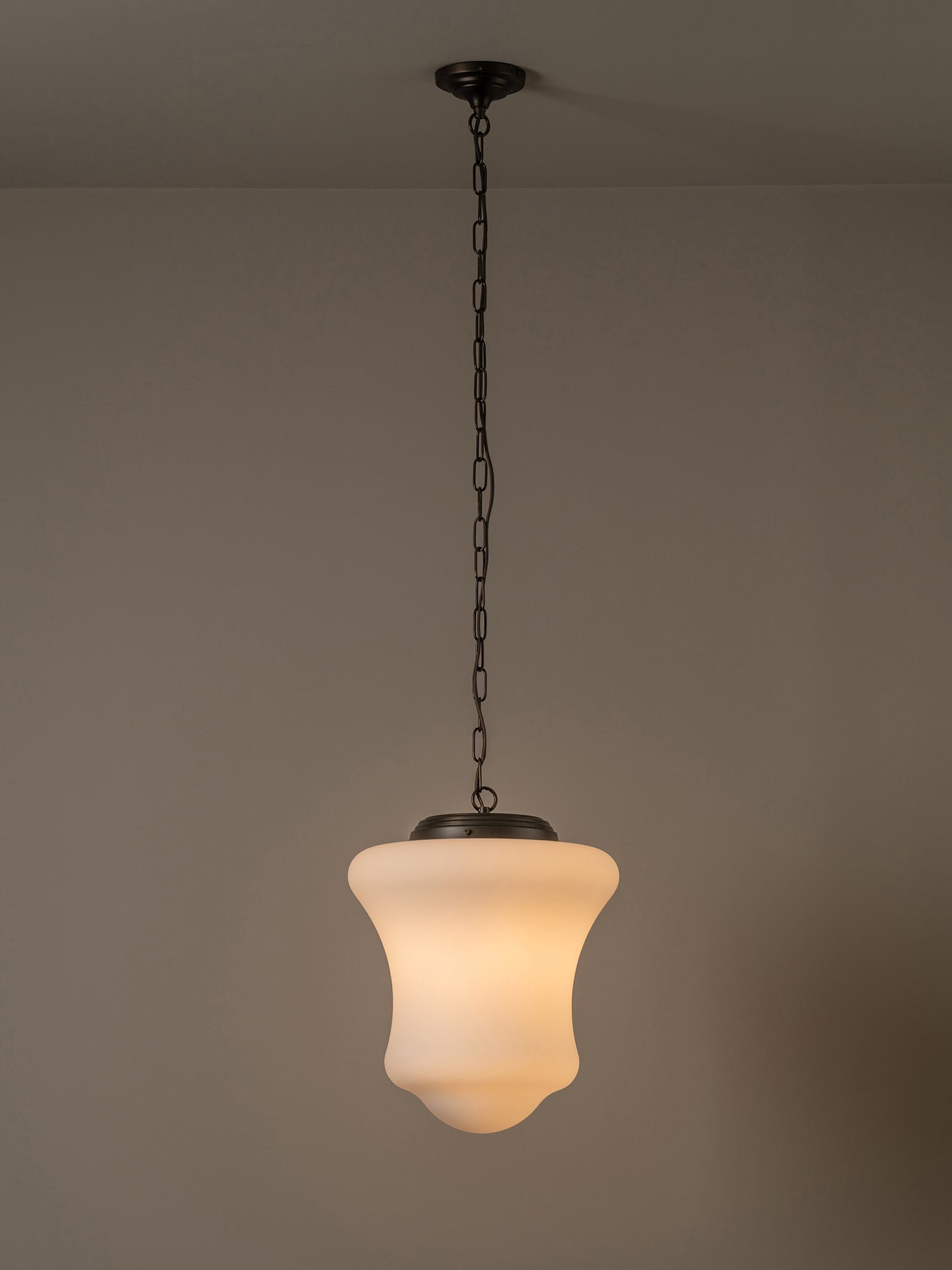 A modern pendant light with a white bell-shaped glass shade and a dark bronze metal canopy and chain, shown hanging against a neutral beige wall.