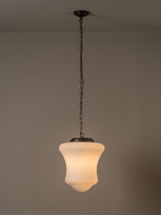 A modern pendant light with a white bell-shaped glass shade and a dark bronze metal canopy and chain, shown hanging against a neutral beige wall.