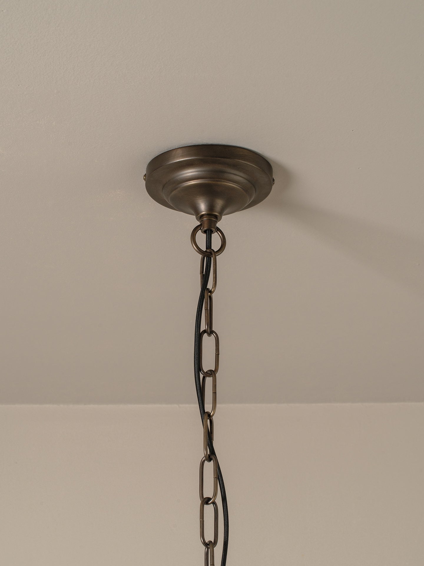 An antique bronze ceiling light fixture canopy hanging from a black metal chain against a neutral off-white wall and ceiling.