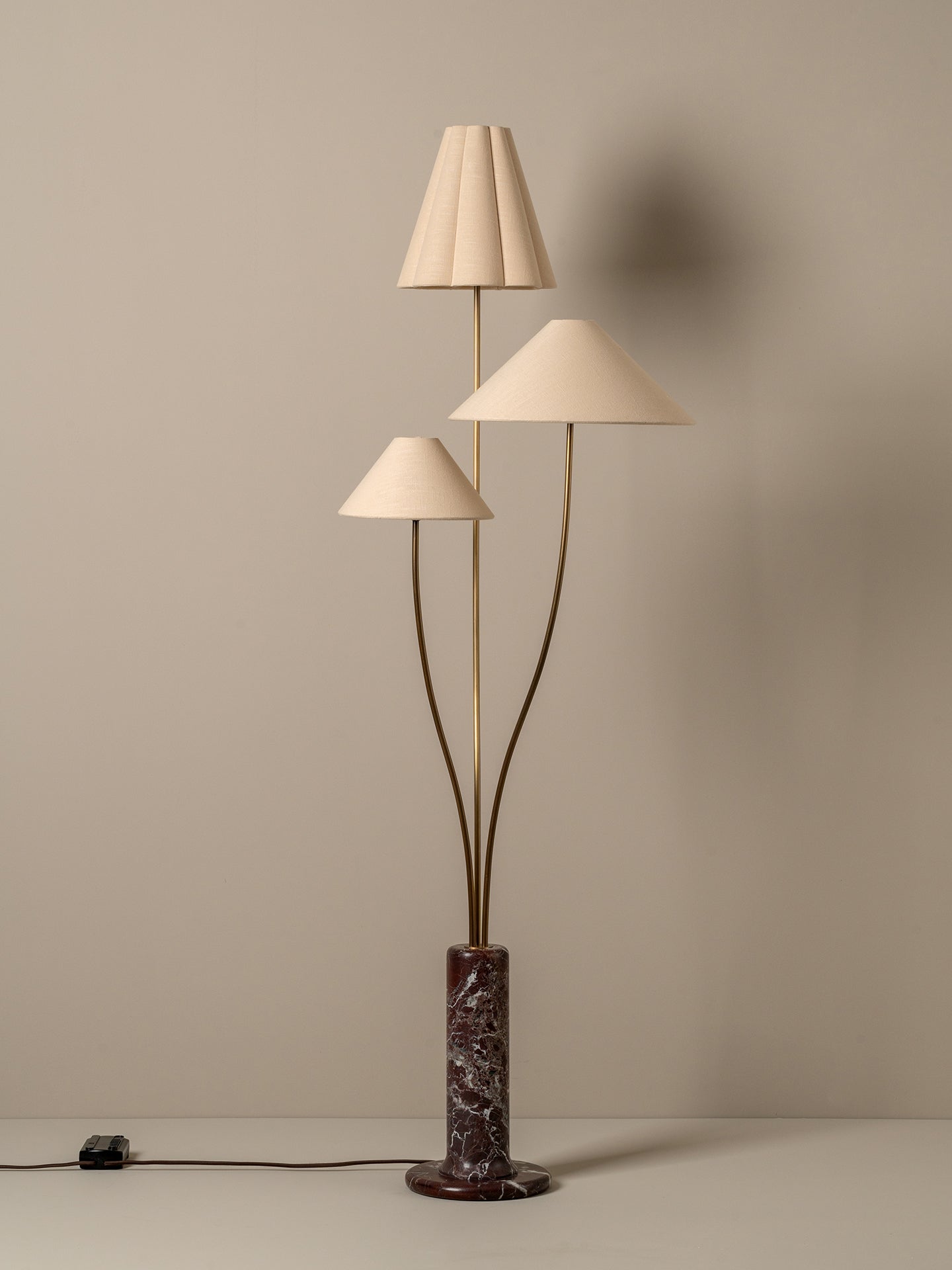 A mid-century modern three-light floor lamp with pleated and conical cream fabric shades, a bronze-finished branching stem structure, and a dark brown and black marble base, set against a warm beige wall.