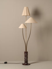 A mid-century modern three-light floor lamp with pleated and conical cream fabric shades, a bronze-finished branching stem structure, and a dark brown and black marble base, set against a warm beige wall.