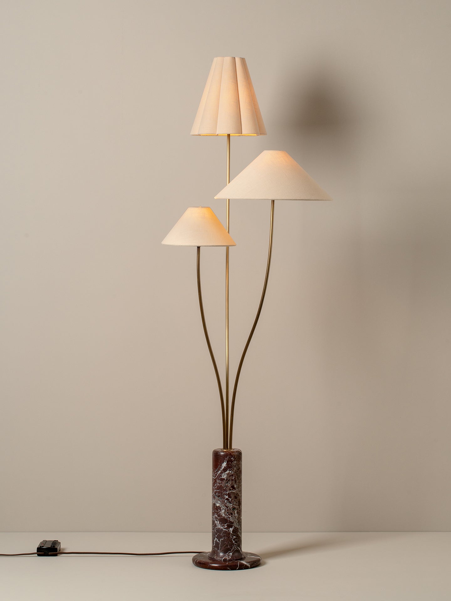 A mid-century modern floor lamp with three tapered cream fabric shades on curved brass stems, mounted on a cylindrical dark brown and black marble base, set against a neutral beige wall.