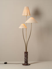 A mid-century modern floor lamp with three tapered cream fabric shades on curved brass stems, mounted on a cylindrical dark brown and black marble base, set against a neutral beige wall.