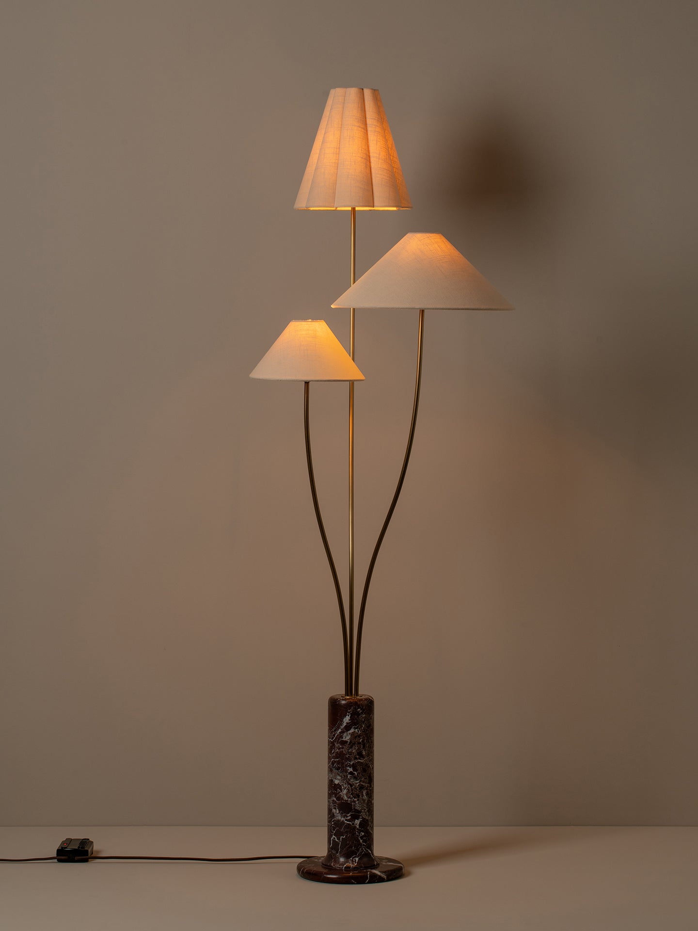 A three-light floor lamp with pleated white fabric shades, dark metal stems, and a dark marble base, shown lit against a plain warm gray wall.