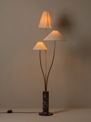 A three-light floor lamp with pleated white fabric shades, dark metal stems, and a dark marble base, shown lit against a plain warm gray wall.