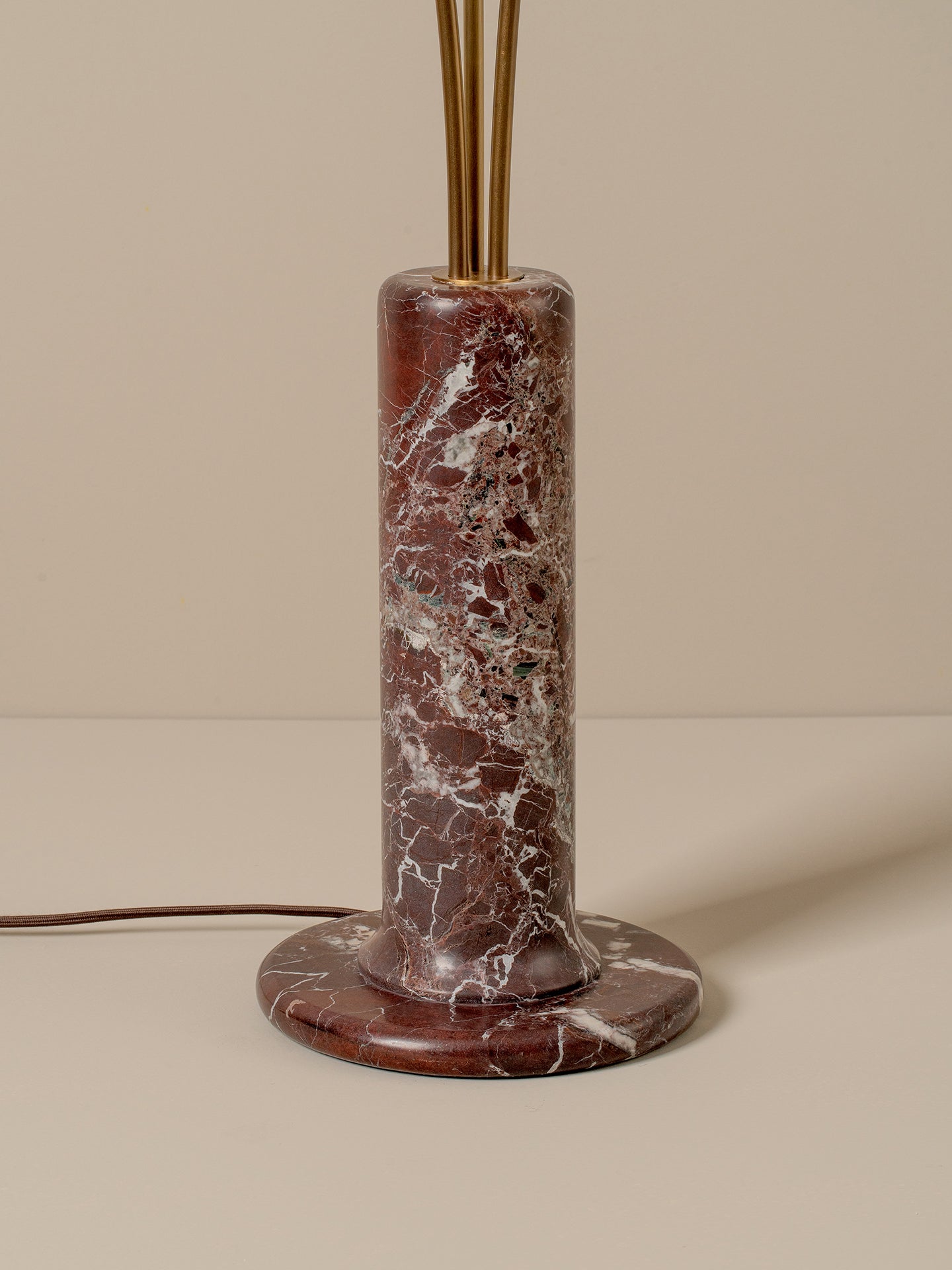 A modern cylindrical table lamp with a round base and central column made of reddish-brown marble with white veining, topped with gold-toned metal rods, set against a warm beige background.