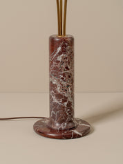 A modern cylindrical table lamp with a round base and central column made of reddish-brown marble with white veining, topped with gold-toned metal rods, set against a warm beige background.