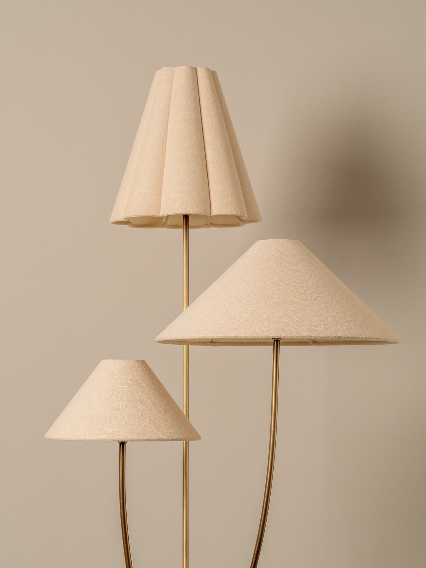 A three-light floor lamp with tapered beige fabric shades and slim gold metal stems, set against a solid warm beige background.