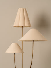 A three-light floor lamp with tapered beige fabric shades and slim gold metal stems, set against a solid warm beige background.