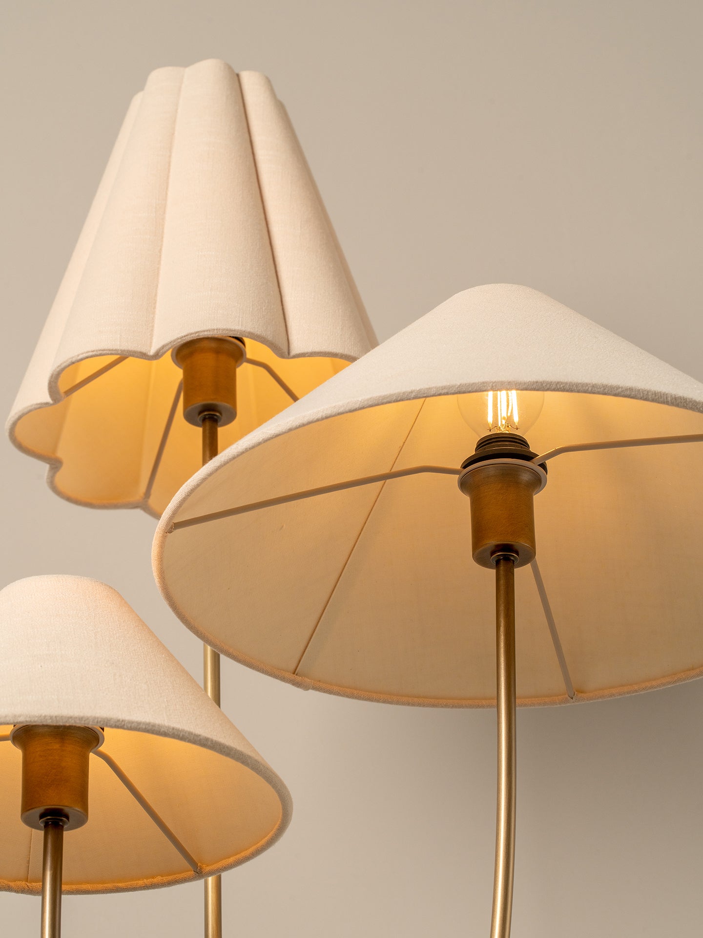 Three modern table lamps with pleated beige fabric shades and warm brass metal stems, shown against a neutral light gray background.