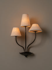 A three-light wall sconce with a dark metal frame and textured beige fabric shades, shown illuminated against a warm taupe wall.