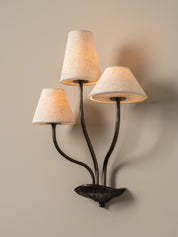 A three-light wall sconce with a dark metal leaf-shaped base and three tapered beige fabric shades, shown lit against a warm neutral wall.