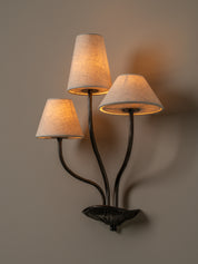 A three-light wall sconce with a dark metal sculptural base and curved arms, each holding a tapered beige linen shade that glows warm yellow when lit, mounted on a plain taupe wall.