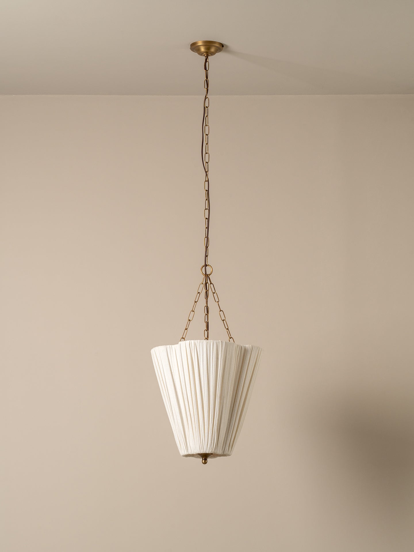 A white pleated conical pendant light hanging from a brass chain and ceiling canopy against a plain beige wall.