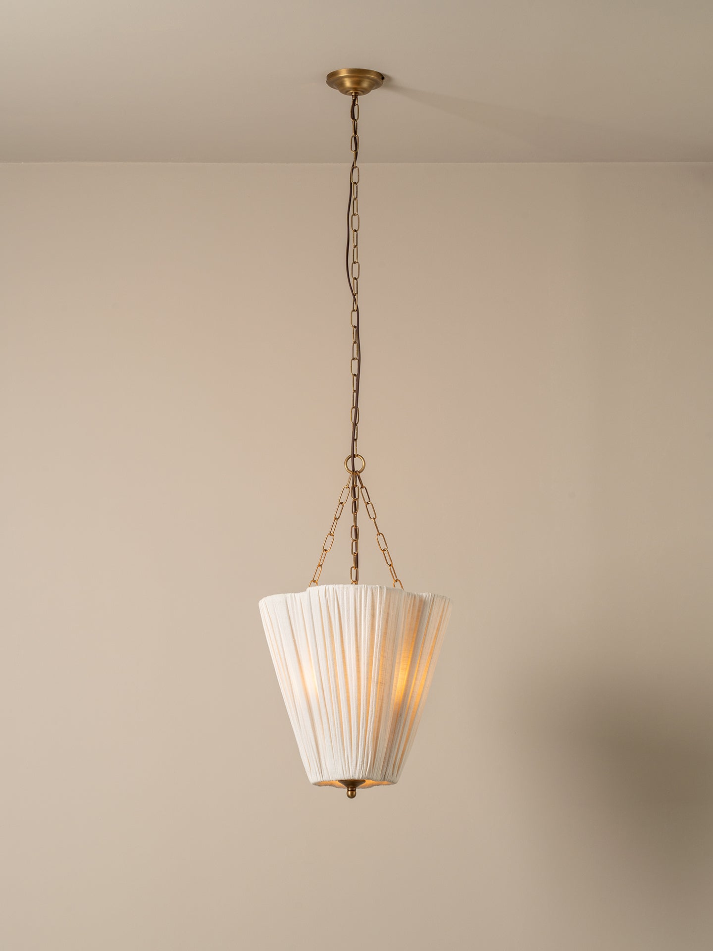 A vintage-style pendant light with a white pleated fabric shade and antique brass hardware, hanging from a ceiling against a plain beige wall.