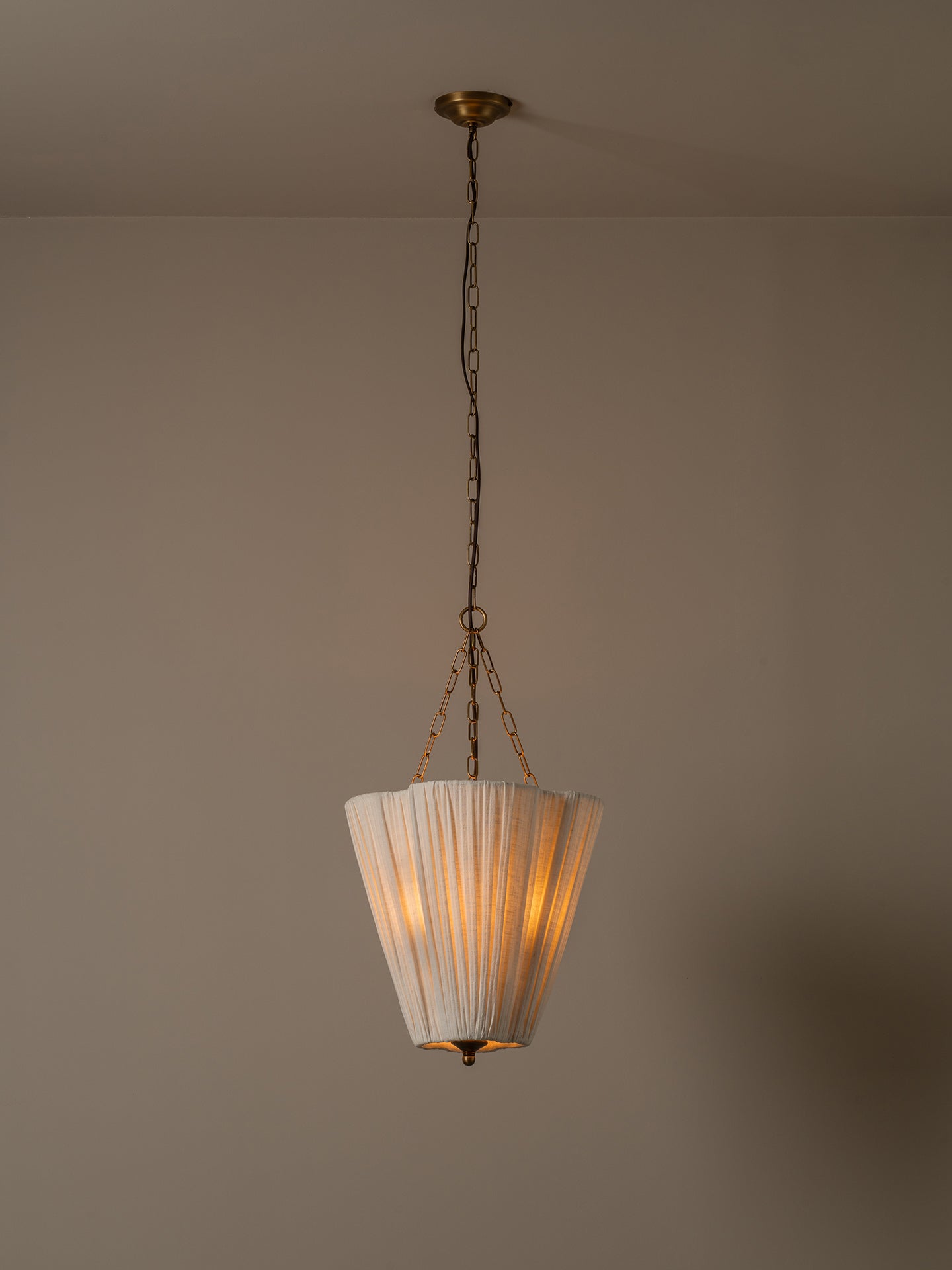 A vintage-style pendant light with a pleated white shade and brass-toned metal hardware, hanging from a chain against a plain beige wall and ceiling.