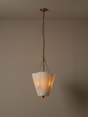 A vintage-style pendant light with a pleated white shade and brass-toned metal hardware, hanging from a chain against a plain beige wall and ceiling.