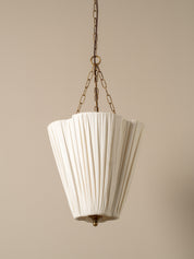A vintage-style pendant light with a pleated white fabric shade and antique bronze metal hardware, suspended from a chain against a solid beige background.