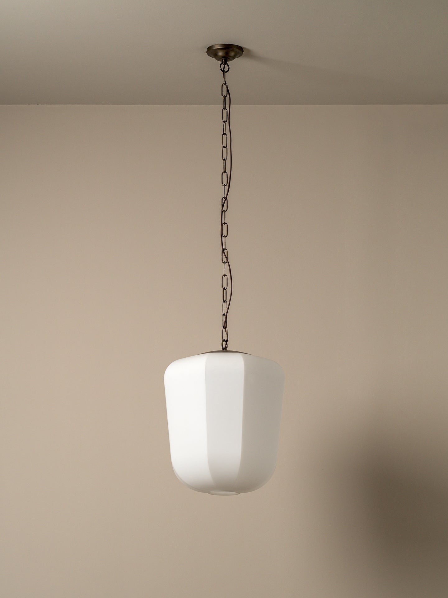 A modern pendant light with a white faceted glass shade suspended by a dark metal chain from a ceiling mount, set against a plain beige wall.