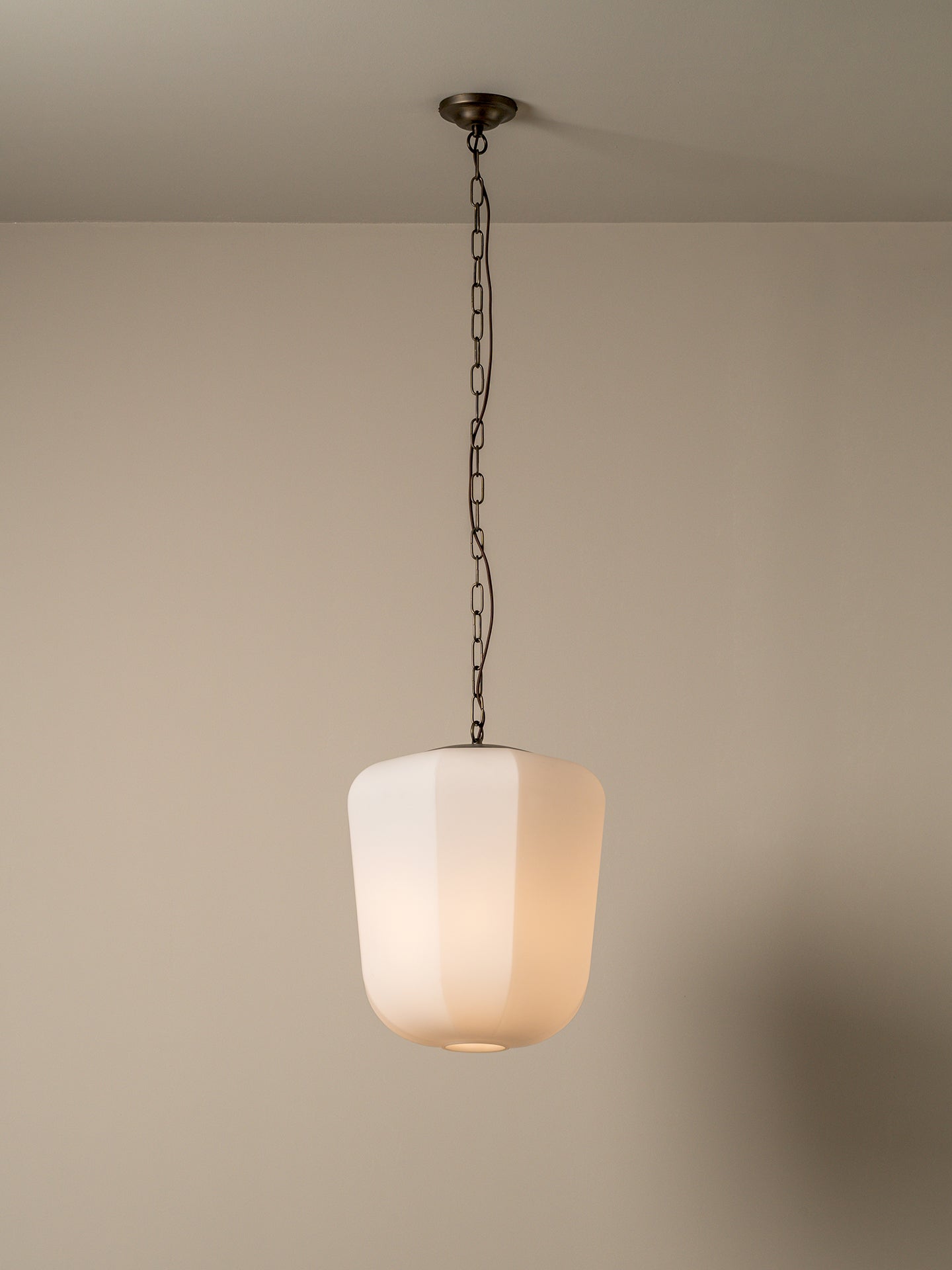 A modern pendant light with a frosted white cylindrical shade and a dark metal chain hanging from a ceiling, shown against a plain beige wall.
