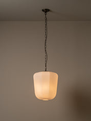 A modern pendant light with a frosted cylindrical glass shade glowing with warm light, suspended by a dark metal chain from a ceiling mount against a neutral beige wall.