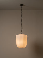 A modern pendant light with a frosted cylindrical glass shade glowing with warm light, suspended by a dark metal chain from a ceiling mount against a neutral beige wall.